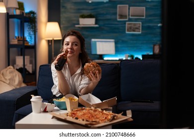 Caucasian Woman Drinking Craft Bottled Beer Eating Delicous Takeaway Hamburger Sitting On Couch In Living Room. Person Laughing Watching Television Having Takeout Order Pizza And Junk Food.