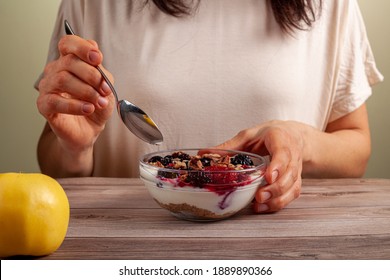 A Caucasian Woman Is Eating A Fresh Home Made Glass Bowl Of Creamy Yogurt Parfait With Berries, Muesli And Seeds In It On Wooden Table Close Up Photo Shows Hand Holding A Spoon With An Apple On Side.