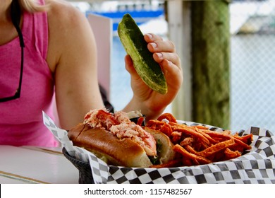 Caucasian Woman Eating Pickle With Lobster Roll And Fries In Basket At Pier Restaurant With Wooden Pier In Background