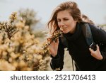 Caucasian woman enjoying nature, smelling yellow flowers. Woman with backpack, walking or hiking outdoors with nature and yellow flowers. Female hiker, nature lover woman, smelling flowers in field.