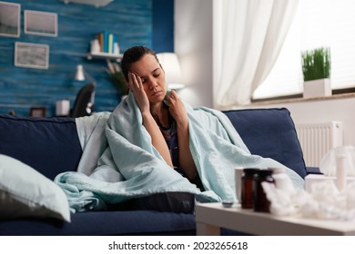 Caucasian woman feeling sick at home with headache, taking medical treatment. Adult in pijamas having cold flu illness sitting on sofa tired, with migraine and sore throat blowing nose
