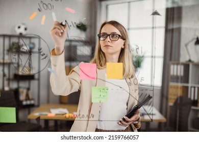 Caucasian woman in glasses holding digital tablet and drawing financial graphic on glass wall before office meeting. Female administrative manager preparing for presentation at work. - Powered by Shutterstock