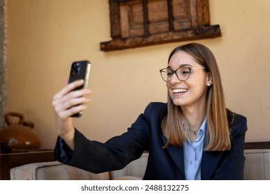 A Caucasian woman with glasses smiling while taking a selfie with her smartphone. She is in an outdoor setting, appearing happy and engaged. - Powered by Shutterstock