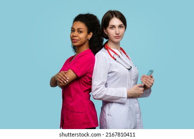 Caucasian Woman GP With Digital Tablet And Dark Skinned Female Doctor Assistant Intern Stand Back To Back On Blue Studio Wall. Multi-ethnic Medical Team Of Healthcare Workers Women Medical Employee