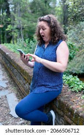 Caucasian Woman, Hair Pulled Back With A Pair Of Sunglasses And A Surprised Face Looking At The Smartphone In A Forest While Resting Leaning Against It. .