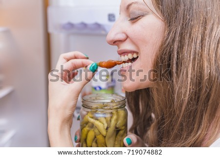 Caucasian woman having a guilty pleasure taste of a chilly pepper, stealing a bite from a fridge