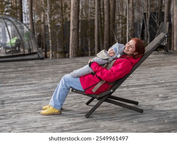 Caucasian woman with her son in an ergo backpack sitting in a wooden deck chair.  - Powered by Shutterstock