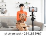 Caucasian woman in her twenties unboxing smartphone on camera. Dressed in orange sweater and brown overalls, she appears lively and engaged. Laptop visible on sofa, with smartphone on tripod.