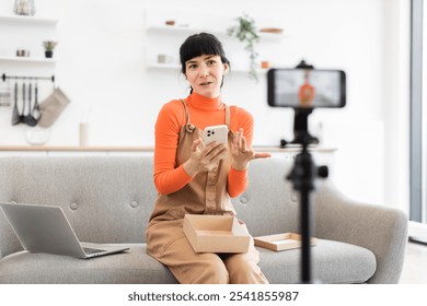 Caucasian woman in her twenties unboxing smartphone on camera. Dressed in orange sweater and brown overalls, she appears lively and engaged. Laptop visible on sofa, with smartphone on tripod. - Powered by Shutterstock