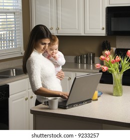 Caucasian Woman Holding Baby  And Typing On Laptop Computer In Kitchen.