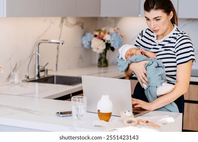 Caucasian woman holding newborn while working on laptop in modern kitchen. Mother multitasking, balance between parenting and remote work. Everyday routine, and technology's role in family life. - Powered by Shutterstock