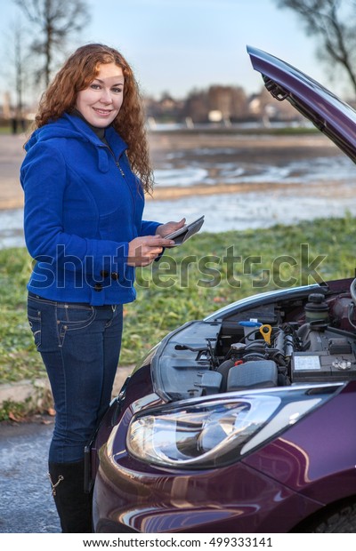 Caucasian woman holding tablet pad and\
standing near broken car with open engine\
hood