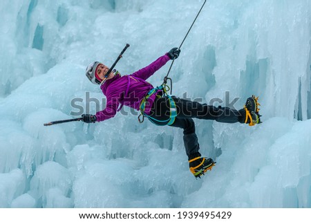Caucasian woman with ice climbing equipment, hiking at a frozen waterfall, holding the ice axe with her mouth and teeth