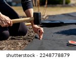 A caucasian woman is kneeling on the ground on a flower bed installing black weed fabric using galvanized steel pegs and a mallet. Concept image for DIY landscaping, weed protection.