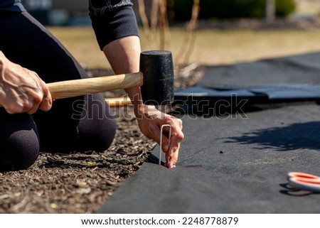 A caucasian woman is kneeling on the ground on a flower bed installing black weed fabric using galvanized steel pegs and a mallet. Concept image for DIY landscaping, weed protection. Foto stock © 
