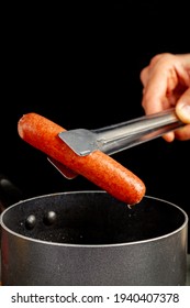 A Caucasian Woman Is Lifting Up A Single Hot Dog Out Of Boiling Water Using A Metal Tong. A Nonstick Cooking Pan And Black Background  Is Seen. Homemade Cooking, Convenient Fast Food Concepts.