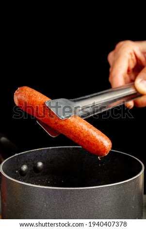 A caucasian woman is lifting up a single hot dog out of boiling water using a metal tong. A nonstick cooking pan and black background  is seen. Homemade cooking, convenient fast food concepts.
