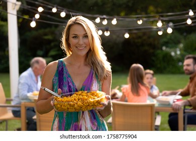 A Caucasian woman with long blonde hair standing in her garden holding a dish of food and smiling to camera, in the background her multi-generation family sitting down at a table for a meal outside - Powered by Shutterstock