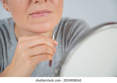 Caucasian woman looks in the mirror and removes hair on her chin herself with the help of tweezers. Hirsutism.