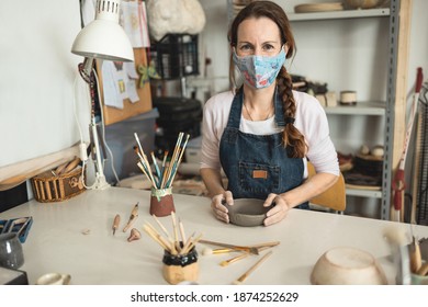 Caucasian woman making ceramic soup plate while wearing protective face mask for coronavirus prevention - Focus on face - Powered by Shutterstock