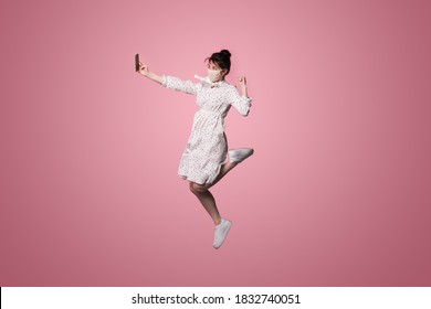 Caucasian woman with a medical mask is wearing a dress and making selfie using a phone while jumping on a pink studio wall