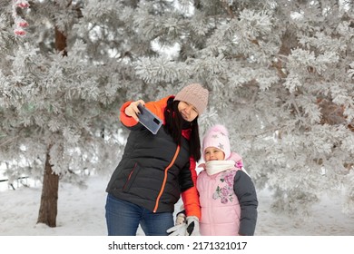 Caucasian Woman Of Middle Age And Little Daughter Making Selfie Smiling Standing On Snowy Ground With Trees In Background Both Looking At Phone. Having Fun Outside In Winter. Family Time.