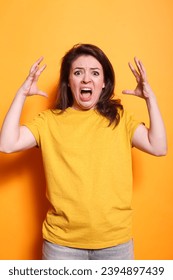 Caucasian woman with open mouth and raised arms, conveying anger and negativity. Annoyed lady in casual clothing stands in studio with orange background, screaming at the camera. - Shutterstock ID 2394897439