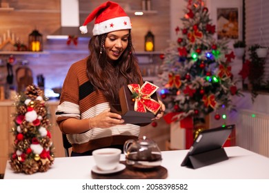 Caucasian Woman Opening Gift Box While Using Video Call Technology With Friends On Tablet. Festive Young Adult With Sweater Receiving Present Feeling Joyful In Christmas Decorated Kitchen