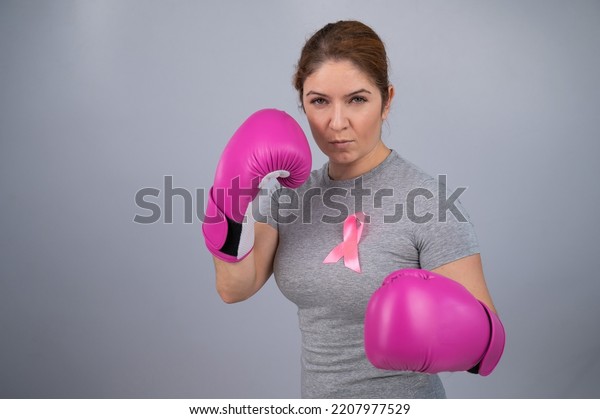 Caucasian
woman in pink boxing gloves with a pink ribbon on her chest on a
gray background. Fight against breast cancer.
