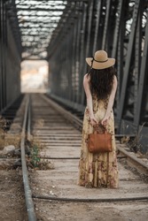 Caucasian Woman Posing With Her Back Turned, Wearing A Long Flowered Vintage Dress, Hat And Handbag, On The Old Railway Tracks In An Industrial Area, With The Handbag Behind Her. 