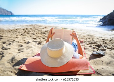 Caucasian woman is reading a book on a sandy beach wearing a straw hat