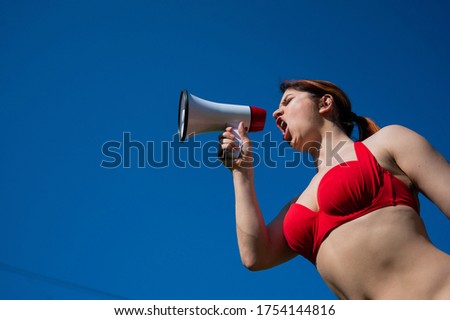 Caucasian woman in red bikini works as a lifeguard on the beach and shouts through a megaphone against a blue sky. A girl in a swimsuit is standing with a loudspeaker. Summer vacation.