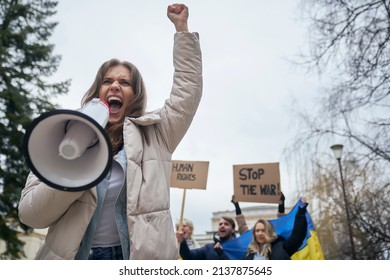 Caucasian woman  screaming through megaphone in front and group of young people manifesting against Ukrainian war in the background - Shutterstock ID 2137875645
