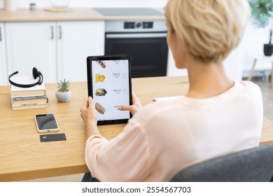 Caucasian woman seated at wooden table ordering food online using digital tablet. Modern kitchen setting with casual feel emphasizes convenience of technology. - Powered by Shutterstock