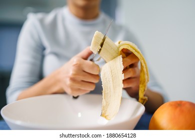 Caucasian woman sitting by the table at home cutting banana - Adult girl female preparing fruit salad at home - healthy eating concept copy space close up