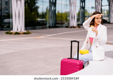 Caucasian woman sitting outside modern building with pink suitcase, yellow lunchbox. Female shields eyes from sun while wearing stylish white blazer and casual attire. - Powered by Shutterstock