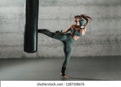 Caucasian woman in sportswear and with boxing gloves kicking bag in the gym. Full length. Wall in background.