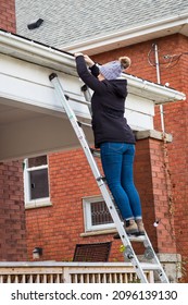 Caucasian Woman Standing On Ladder Putting Up Christmas Lights On Roof Of Red Brick Home. 