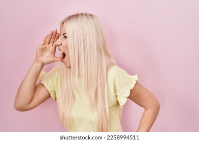Caucasian woman standing over pink background shouting and screaming loud to side with hand on mouth. communication concept.  - Shutterstock ID 2258994511