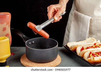 A caucasian woman is taking a fresh boiled hot dog sausage from a pot  using metal tong in order to place it into a hot dog bun. Hot pot is on a cork trivet on stone couter. Ketchup and mustard nearby - Powered by Shutterstock