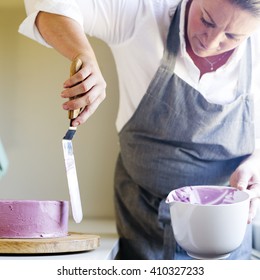 Caucasian Woman Trimming The Cream Icing On A Lilac Cake With A Spatula