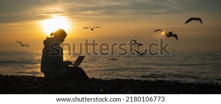 Caucasian woman typing on a laptop on the seashore at sunset. Freelance work. Wide screen. 