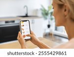 Caucasian woman uses smartphone app to order food online from home kitchen setting. Image captures modern and convenient lifestyle of digital food delivery.