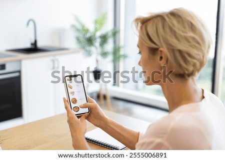 Caucasian woman using smartphone to order food online. Blonde adult sitting at home kitchen browsing through delivery options with phone. User-friendly experience of digital food-ordering platform.
