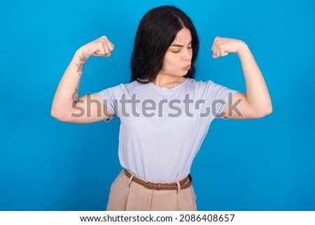 Caucasian woman wearing blue T-shirt over blue background showing arms muscles smiling proud. Fitness concept.