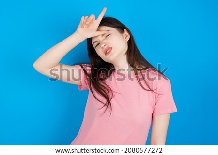 Caucasian woman wearing pink T-shirt over blue background making fun of people with fingers on forehead doing loser gesture mocking and insulting.