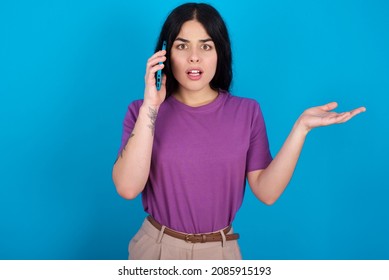 Caucasian Woman Wearing Purple T-shirt Over Blue Background Talking On The Phone Stressed With Hand On Face, Shocked With Shame And Surprise Face, Angry And Frustrated. Fear And Upset For Mistake.