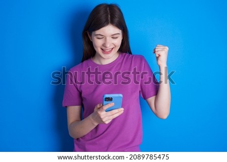Caucasian woman wearing purple T-shirt isolated over blue background holding in hands cell and rising his fist up being excited after reading good news.