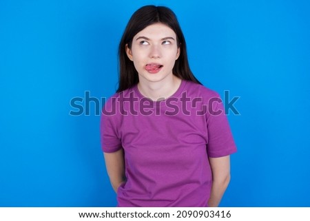 Caucasian woman wearing purple T-shirt isolated over blue background showing grimace face crossing eyes and showing tongue. Being funny and crazy