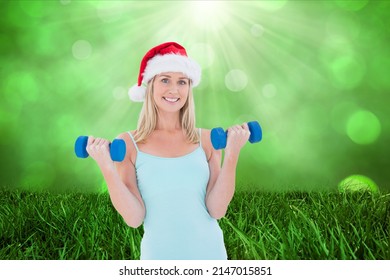 Caucasian woman wearing santa hat holding dumbbells against grass and spots of light with copy space. chritmas and fitness concept